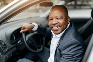 Portrait of confident black businessman smiling while sitting in car, hands on wheel. African...
