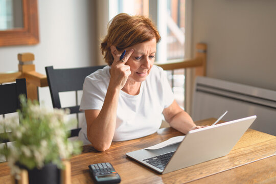 Sad frustrated senior woman pensioner having depressed look, holding hand on her face, calculating family budget, sitting at kitchen counter with laptop, papers, coffee, calculator and cell phone
