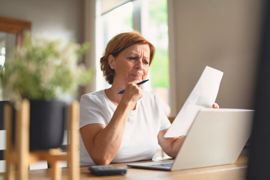 Sad frustrated senior woman pensioner having depressed look, holding hand on her face, calculating family budget, sitting at kitchen counter with laptop, papers, coffee, calculator and cell phone