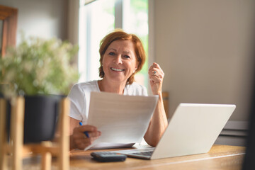happy senior woman calculating family budget, sitting at kitchen counter with laptop, papers,