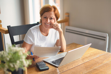 happy senior woman calculating family budget, sitting at kitchen counter with laptop, papers,