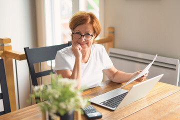 happy senior woman calculating family budget, sitting at kitchen counter with laptop, papers,