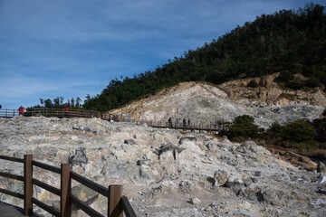 Banjarnegara, Indonesia-July 31, 2022: Tourists on wooden pathways observing the steaming Kawah Sikidang volcanic crater in Dieng, surrounded by rugged terrain and green trees under a blue sky.