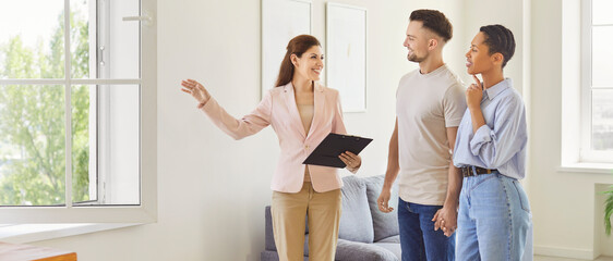 Female realtor showing house to a young happy couple looking at the view from the window....