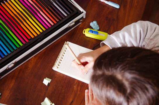 Top view of a young girl drawing on a notebook sitting at a wooden table with a collection of colorful pencils. 