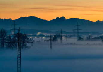 Aerial view of mist-shrouded fields and towering power lines beneath a soft, orange-tinged sky, with mountains in the distance, Marktoberdorf, Bayern, Germany.