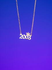 Close-up of a metallic gold pendant in the shape of "2003" on a chain, on a  purple background