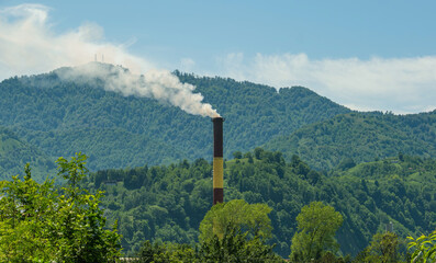 Industrial chimney emitting smoke in mountain landscape. Tall industrial chimney releases white smoke into the air against a backdrop of green forested mountains. Concept of environmental impact.