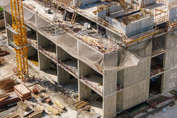 Concrete building under construction featuring multiple floors, yellow tower crane and safety nets installed around the edges. Various construction materials are scattered on the ground below.