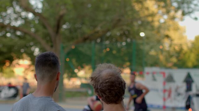 Two young men watch an exhilarating sports game taking place at a lively park during the beautiful sunset, surrounded by lush greenery and various enjoyable outdoor activities and people