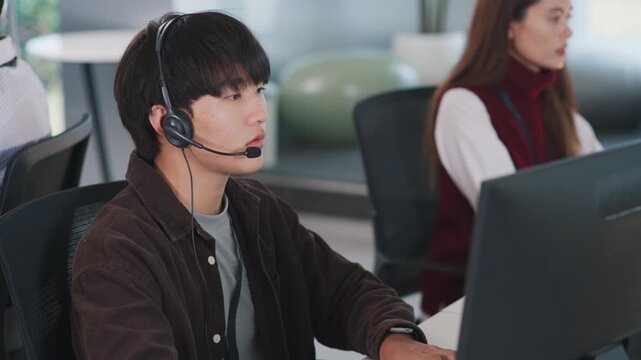 Young man wearing headset working at computer in business meeting tech support call center with focused expression and female colleague in office customer support environment - Powered by Adobe