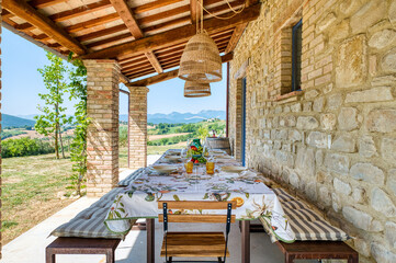 A rustic stone and wood portico with a table set outdoors, featuring a floral tablecloth, glasses, fresh fruit, and wicker lamps. Natural Mediterranean light enhances the Italian villa and green hills