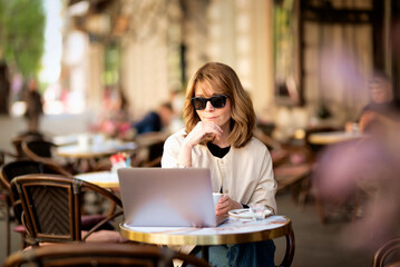 Middle aged woman sitting at sidewalk cafe in the city and using laptop
