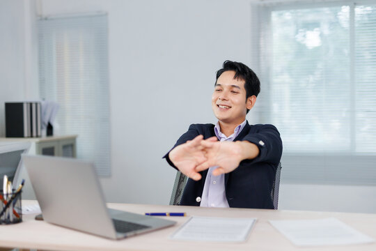 Asian businessman stretching his arms at an office desk, enjoying a well deserved break from work while seated with a laptop
