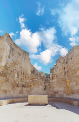 The ruins of an ancient stone structure with high, curved walls and a central pedestal under a bright blue sky with white clouds.
