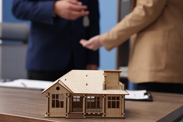 Real estate agent giving key to client at desk in office, focus on house model