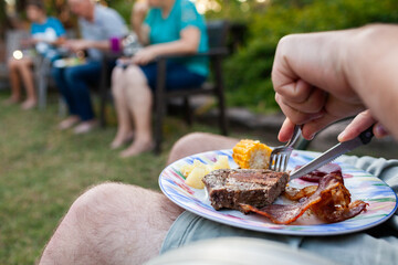 man cutting into steak on plate with knife and fork at outdoor family bbq event