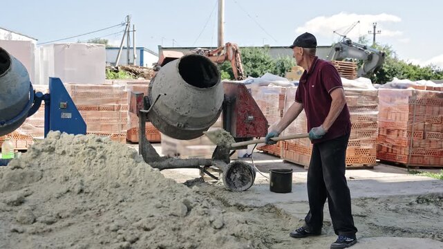 Construction worker mixing sand near cement mixer. Older man at construction site using a shovel to mix sand in front of a concrete mixer, with bricks and machinery in background.