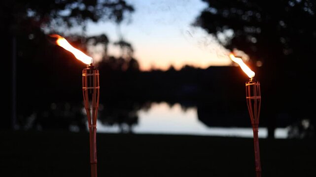 A row of tiki torches casting warm flames and smoke trails against a backdrop of silhouetted trees and twilight sky, creating a moody outdoor ambiance