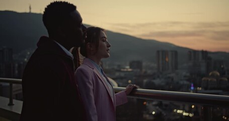 Two business executives discuss strategy on a rooftop at sunset, overlooking a cityscape. They point and gesture, enjoying the view and their success. - Powered by Adobe
