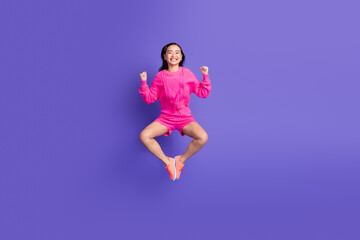 Young woman in vibrant pink jumper jumping joyfully against a purple background, expressing charm and positive emotion