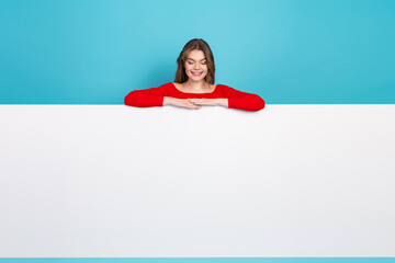 Young woman in bright red outfit leaning on a large blank sign with cheerful expression against a...