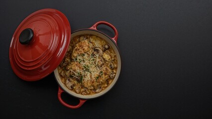 Top-down photo of a baked chicken and mushroom dish with cheese in a ceramic pot, black background, empty space for text