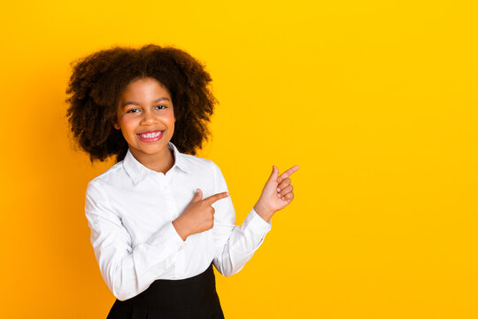 Smiling young girl pointing with both hands toward copy space on yellow background in stylish school uniform