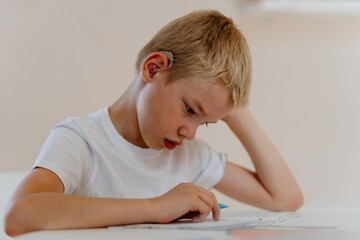 Naklejka premium A boy with hearing aids concentrates on homework at a sunlit desk, embodying joyful learning and resilience. Authentic moment of inclusive education and childhood determination
