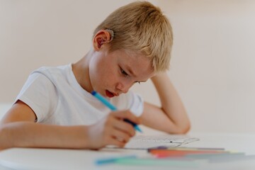 A boy with hearing aids concentrates on homework at a sunlit desk, embodying joyful learning and...