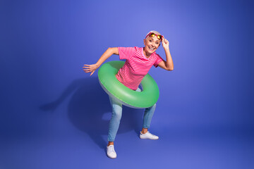 Obraz premium Joyful mature woman in casual attire with green swimming ring on violet background, showcasing a playful summer vibe