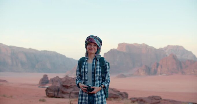 Young Woman Tourist in turban smiles at the camera holding an analog photo camera in Wadi Rum desert in Jordan. Portrait of adventurous journalist and cultural explorer. 4K medium orbit shot