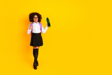 Happy schoolgirl wearing glasses and backpack holding bottle, posing on yellow background in formal school uniform