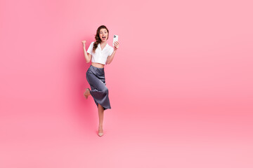 Joyful young woman celebrating with a smartphone against a lively pink background in stylish casual...
