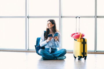 Passenger woman sitting cross-legged in an airport terminal holding a smartphone and passport with...