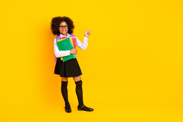 Smiling young schoolgirl holding notebooks and pointing on yellow background representing education...