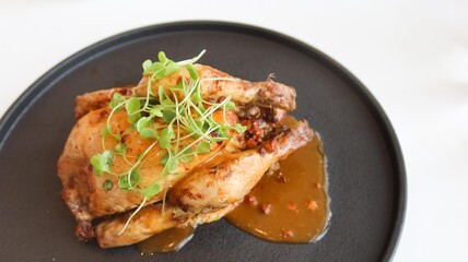 close up of dish of roasted golden-brown chicken with crispy skin.  Garnished with fresh microgreens on top and with savory sauce with copy space