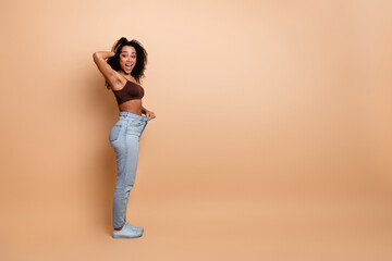 Young woman celebrating weight loss in oversized jeans on a beige background, promoting fitness and...