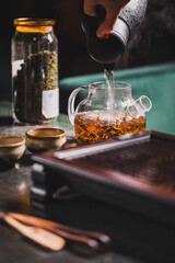 Glass teapot with tea leaves being filled with hot water from a kettle on a wooden tray, surrounded by cups and a jar of loose tea. Cozy tea preparation scene.