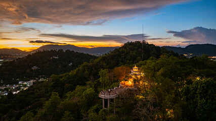 Fototapeta premium A scenic aerial view of a town nestled among lush green mountains at sunset, with a tower structure perched on the hillside and a vibrant orange sky creating a warm, tranquil atmosphere.