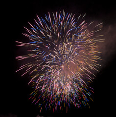 A vibrant, large explosion of multicolored fireworks bursts in the dark night sky, creating a...