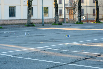 An empty, asphalt urban parking lot with white lines, trees, and buildings in the background, showing some scattered litter on the ground. © tino