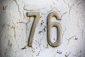 Metal house number 76 mounted on a white wall with peeling paint and rust stains. The textured...