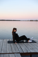 Fototapeta premium Woman practicing yoga on a wooden pier by the water at sunrise. Peaceful atmosphere, healthy lifestyle, balance and harmony with nature. Woman meditating on a yoga mat at wooden pier by the water