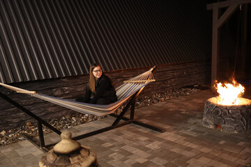 A young woman relaxes on a striped hammock by a cozy firepit at night in a pine forest. Peaceful...