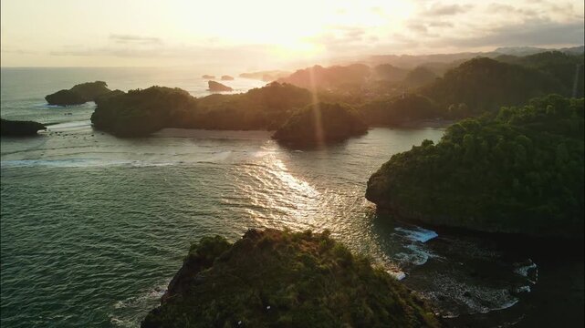A stunning seascape unfolds with numerous green-topped sea stacks and rocky islands scattered across the ocean, all bathed in the warm, hazy light of sunset. Watu Karung Beach, Pacitan.