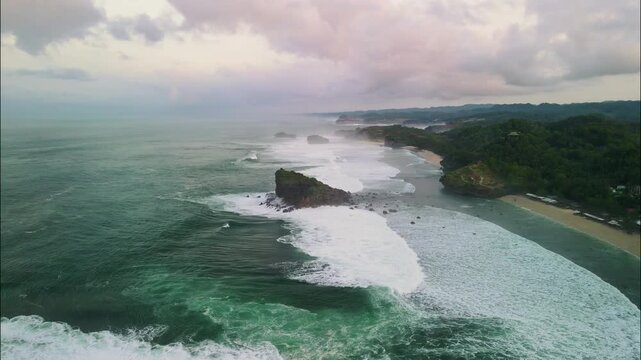 From a cliff-top viewpoint, this stunning aerial shot captures a crescent beach and a bay dotted with numerous green, rocky sea stacks and crashing waves. Aerial Watu Karung beach in Pacitan.
