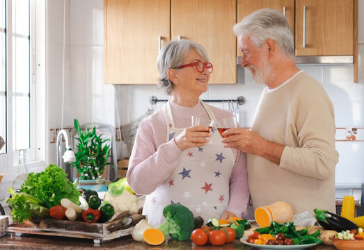 Cheerful senior couple toasting with red wine glasses working together in the home kitchen preparing vegetables for a healthy soup. Joyful elderly people appreciate a vegetarian eating