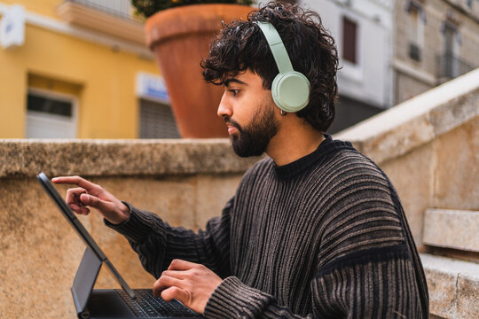 Young digital nomad is working outdoors, using his tablet and headphones in urban city
