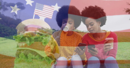 Chatting friends wearing sweaters, jeans sipping cups on grassy field, featuring flag and burger